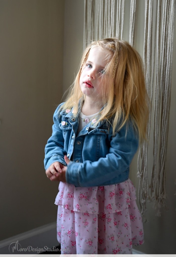 One of the twins in a denim jacket and floral dress, standing by a window