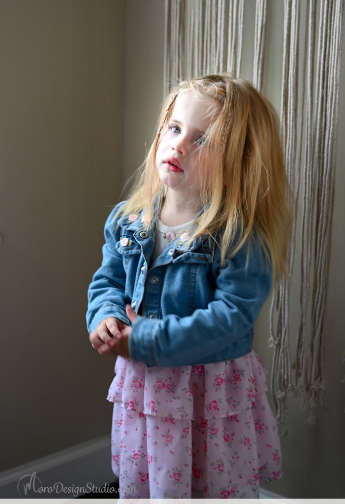 One of the twins in a denim jacket and floral dress, standing by a window