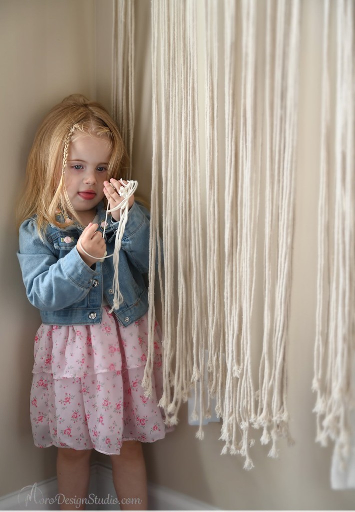 One of the twins holding cream macrame cords, wearing a denim jacket and floral dress