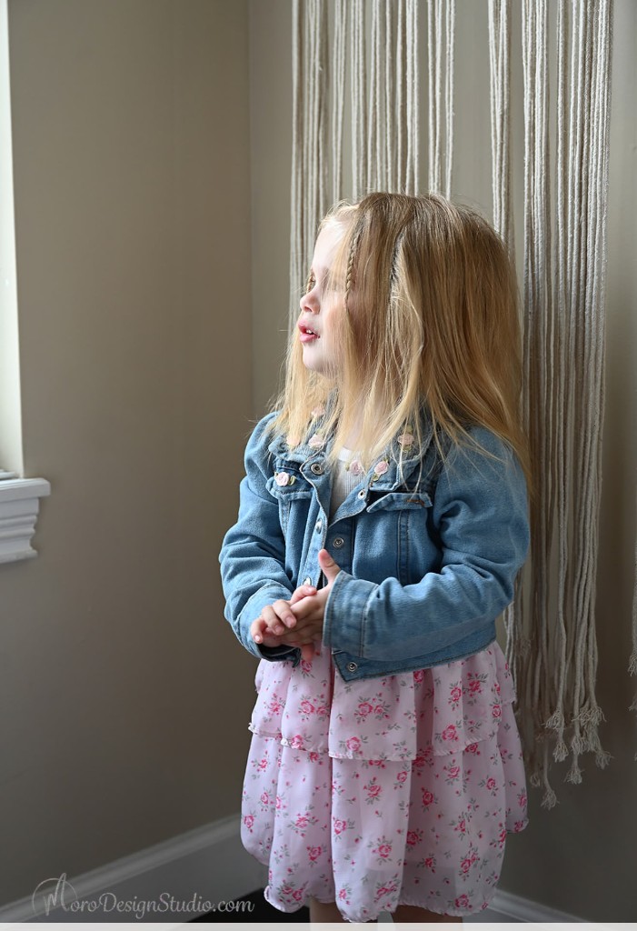 One of the twins standing by a window in a denim jacket and floral dress, looking toward the light